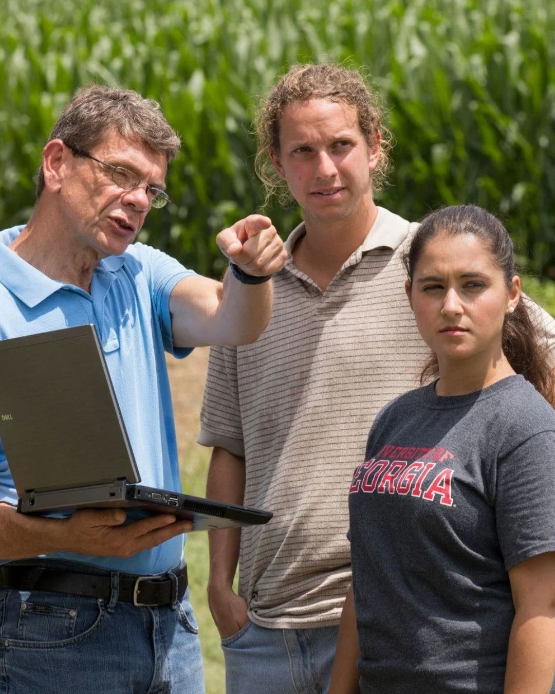 Professor holding a computer instructing two graduate students standing in a field