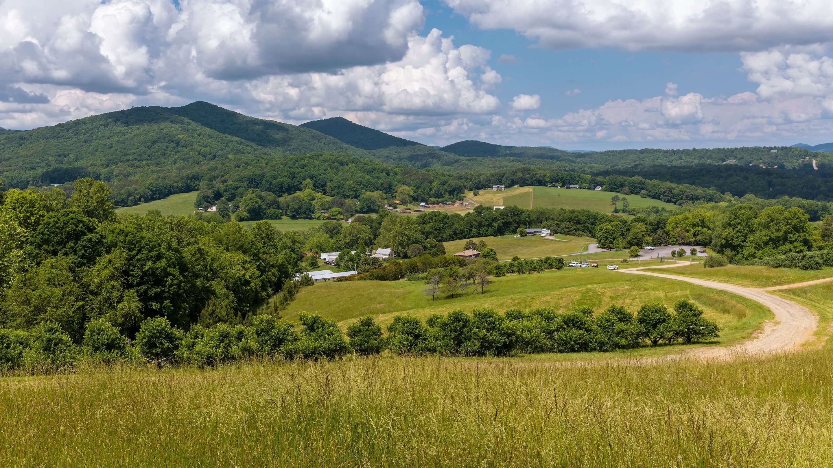 Aerial view of a mountain valley with rolling green pastures, a winding gravel road, scattered farmhouses and barns, and forested hills beneath a partly cloudy sky.